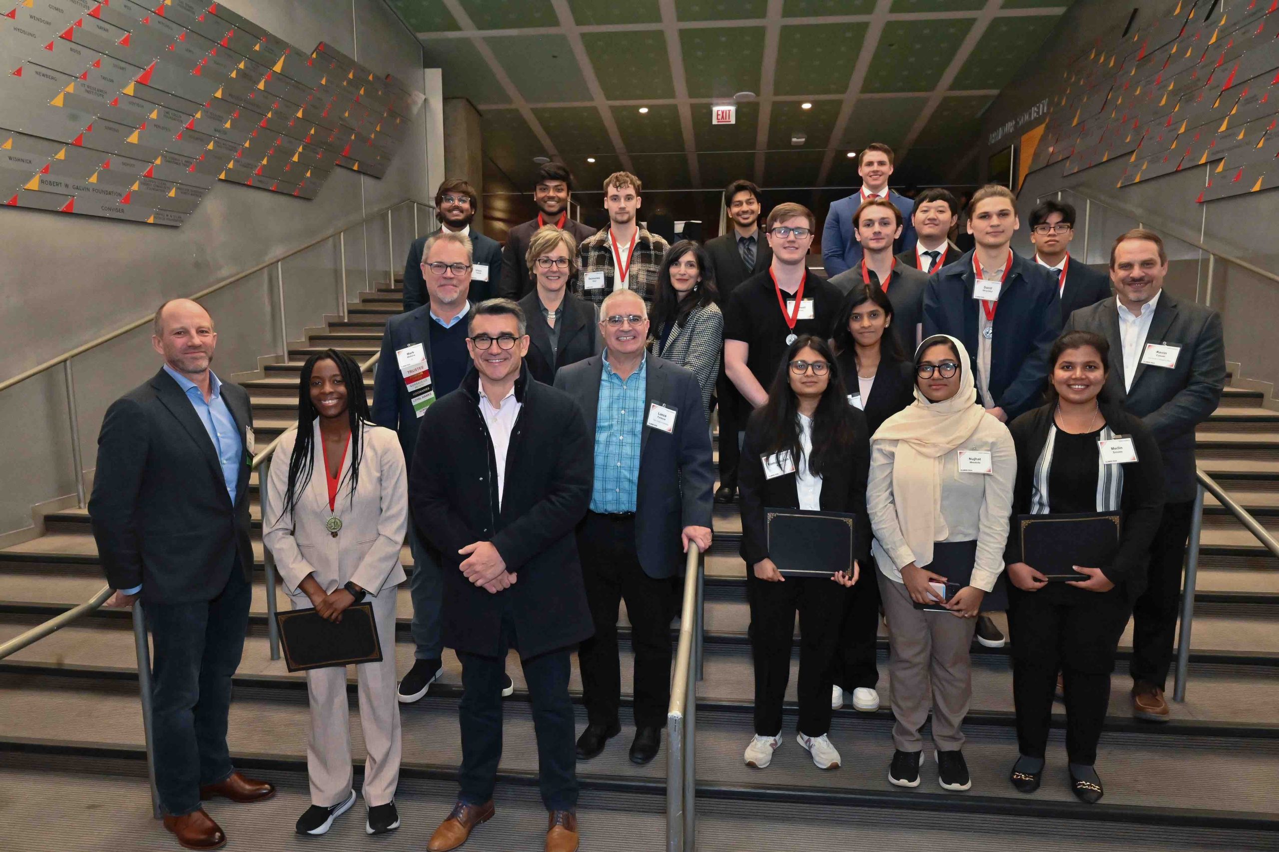 A group photo of the 2024 Grainger Prize participants and event organizers. The group consists of approximately 25 people standing on a staircase. Some individuals wear medals around their necks, while others hold certificates. The participants are dressed in business and formal attire, smiling at the camera. The background features a wall with a design of triangular elements in red, yellow, and orange. The staircase is framed by metal railings on each side.