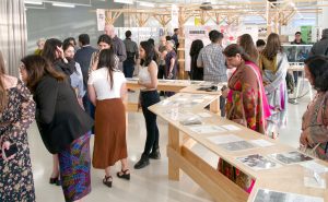 About twenty people stand in a room. Some are talking to each other and others are looking at posters in the back of the room or displays on a long wooden table down the middle of the room.