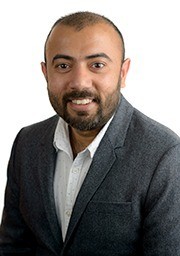 A man wearing a white button-down shirt and a gray blazer smiles for a portrait in front of a white background.