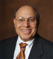 A man wearing round wire glasses, a white shirt, a black pinstripe blazer and brown striped tie smiles in a portrait against a brown background.