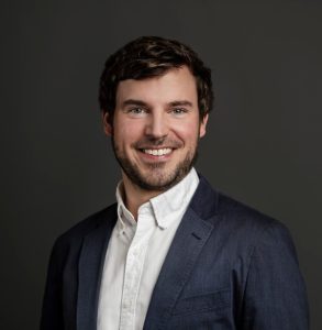 A man wearing a white button-down shirt and dark blue blazer smiles in a portrait in front of a neutral background.