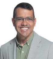 A man wearing half-rim glasses, a green button-down shirt, and gray blazer smiles in a portrait against a white background.