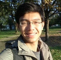 A man wearing wire glasses and a black backpack smiles at the camera. He is outdoors with some trees behind him.